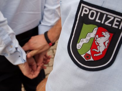 Polizist legt einer Person Handschellen an; das Logo der Polizei NRW ist im Vordergrund